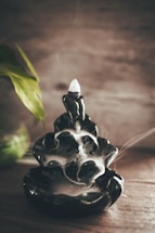 A dark ceramic incense holder with smoke gently flowing down its cascading layers, creating a serene atmosphere. To the left, a green plant leaf adds a touch of nature, contrasting with the earthy tones of the wooden surface.