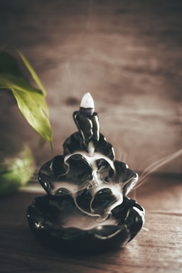 A dark ceramic incense holder with smoke gently flowing down its cascading layers, creating a serene atmosphere. To the left, a green plant leaf adds a touch of nature, contrasting with the earthy tones of the wooden surface.