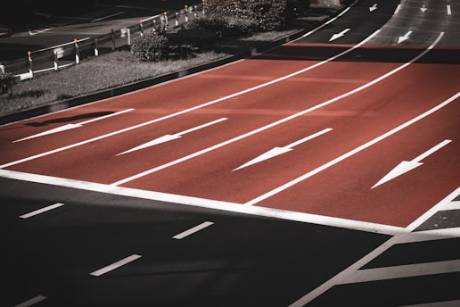 A multi-lane road intersection with red asphalt, featuring several white directional arrows indicating lane usage. The lanes are separated by white lines, and there is greenery and guardrails along the sides.