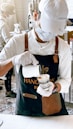 A barista wearing a white cap, mask, and gloves is carefully pouring milk into a coffee cup. The barista is dressed in a white shirt and a dark apron with leather accents. In the background, another person is seen working, and there are stacks of cups and coffee-making equipment on a counter.