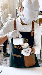 A barista wearing a white cap, mask, and gloves is carefully pouring milk into a coffee cup. The barista is dressed in a white shirt and a dark apron with leather accents. In the background, another person is seen working, and there are stacks of cups and coffee-making equipment on a counter.
