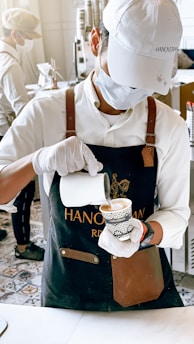 A barista wearing a white cap, mask, and gloves is carefully pouring milk into a coffee cup. The barista is dressed in a white shirt and a dark apron with leather accents. In the background, another person is seen working, and there are stacks of cups and coffee-making equipment on a counter.