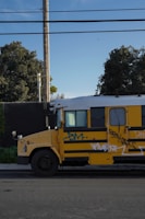 A yellow school bus with graffiti is parked on the side of a street. The bus shows signs of wear and is positioned under clear blue skies. In the background, there are tall trees and utility poles.