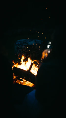 A cozy campfire glowing amber against a deep coal black night, surrounded by worn leather-bound books and a vintage microphone.