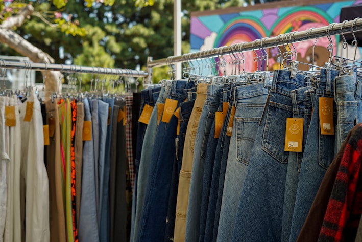 A rack of hanging pants, primarily showcasing denim jeans, with price tags attached. Various colors and styles can be seen, including lighter and darker shades, set against the backdrop of a colorful mural and leafy trees.