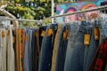 A rack of hanging pants, primarily showcasing denim jeans, with price tags attached. Various colors and styles can be seen, including lighter and darker shades, set against the backdrop of a colorful mural and leafy trees.