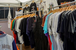 A variety of colorful t-shirts are displayed on wooden hangers under a white canopy. The t-shirts feature different graphics and designs, including one prominently displaying the Mickey Mouse logo. The setting appears to be an outdoor market or pop-up shop.