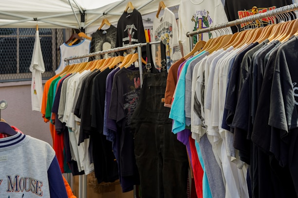 A variety of colorful t-shirts are displayed on wooden hangers under a white canopy. The t-shirts feature different graphics and designs, including one prominently displaying the Mickey Mouse logo. The setting appears to be an outdoor market or pop-up shop.