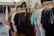A variety of colorful t-shirts are displayed on wooden hangers under a white canopy. The t-shirts feature different graphics and designs, including one prominently displaying the Mickey Mouse logo. The setting appears to be an outdoor market or pop-up shop.