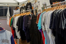 A variety of colorful t-shirts are displayed on wooden hangers under a white canopy. The t-shirts feature different graphics and designs, including one prominently displaying the Mickey Mouse logo. The setting appears to be an outdoor market or pop-up shop.