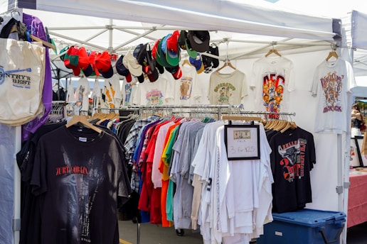 A market stall displaying a collection of vintage t-shirts and hats under a white canopy. The t-shirts are various colors and feature different graphic designs. Several baseball caps are hung along the top frame of the stall. A sign indicates a special offer of three t-shirts for $25. A blue storage bin is visible at the bottom right.
