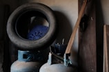 A worn-out tire is placed on top of a blue plastic chair with a decorative pattern. Below them, two gas cylinders are positioned next to a wooden plank leaning against a wall. The setting suggests a storeroom or garage with a rustic and somewhat dimly lit atmosphere.