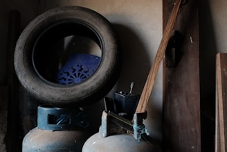 A worn-out tire is placed on top of a blue plastic chair with a decorative pattern. Below them, two gas cylinders are positioned next to a wooden plank leaning against a wall. The setting suggests a storeroom or garage with a rustic and somewhat dimly lit atmosphere.