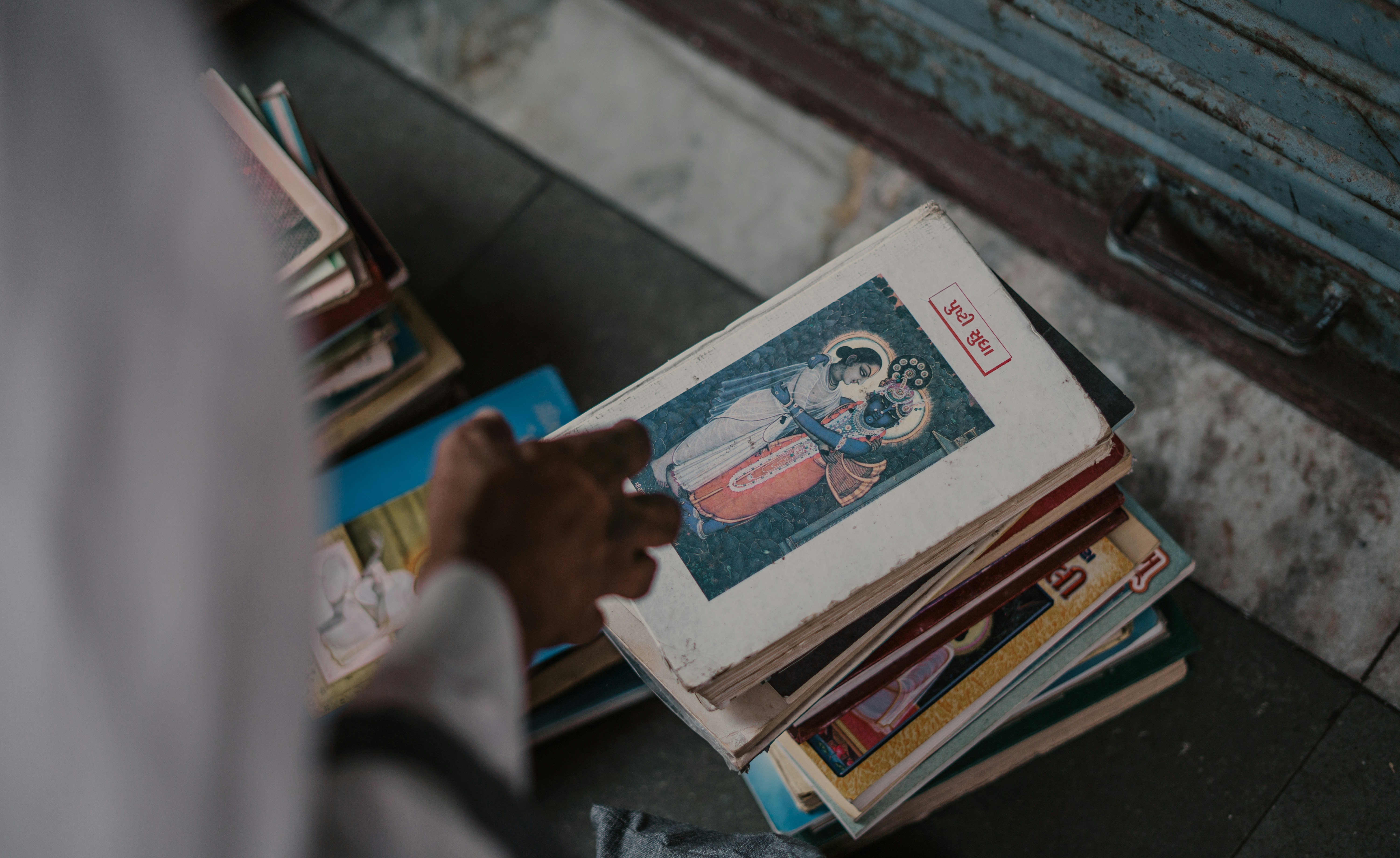 A stack of school textbooks, one open to a page showing lyrics of a Hindi film song, with a student's hand pointing at it.
