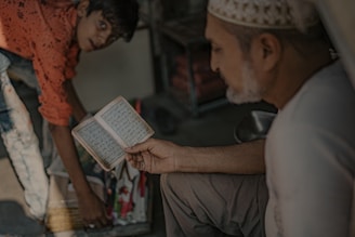 An elderly man is sitting while holding and reading a small open book, presumably a religious text. Beside him, a young boy is bending down, appearing to arrange or interact with items on the ground, including a board. The setting suggests an informal or everyday environment with soft, muted lighting.