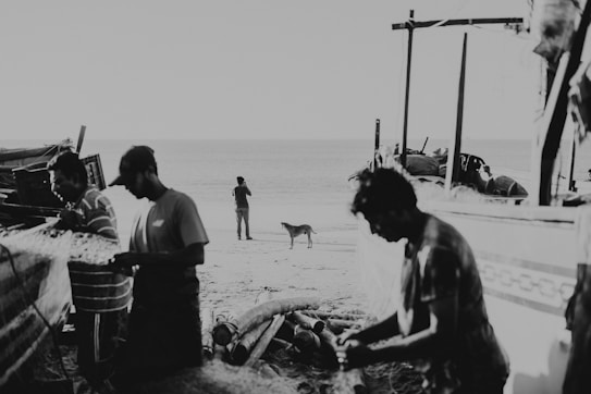 A black and white image featuring people working with fishing nets on a beach. In the background, the ocean is visible with a person standing near the water, and a dog nearby. Boats and fishing equipment are scattered around the area, creating a scene of routine coastal life.