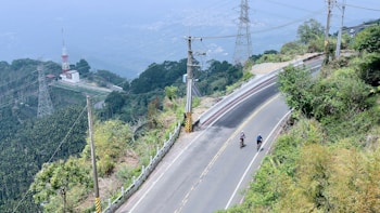 A winding mountain road with two cyclists riding along it, surrounded by lush greenery and steep hills. There are power lines and a transmission tower visible, along with a structure that resembles a small communications station.