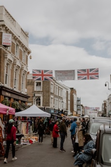 A bustling street market scene featuring multiple stalls with awnings lining the road. People are walking and browsing the shops on Portobello Road, as indicated by a banner overhead. The street is decorated with British flags, and the buildings have a classic architectural style with brick facades.