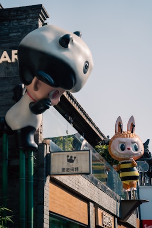 Two large, colorful statues resembling cartoon characters are mounted on the exterior of a building. One character resembles a panda, while the other looks like a rabbit dressed in a bee costume. The building appears to be constructed with gray bricks and features various signs.