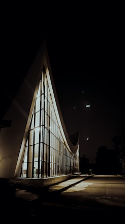 A modern architectural structure with a prominent triangular shape illuminated from within. The walls are composed of large glass panels, creating a striking contrast against the dark night sky. The building's exterior is sleek and minimalistic, casting soft shadows on the surrounding pavement.