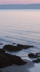 Calm coastal scene at sunset with gentle waves and muted taupe rocks under a soft cream sky.