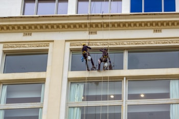 Two workers are suspended on ropes cleaning the windows of a tall building. They are equipped with safety gear, including helmets and harnesses, and are positioned near large glass windows.