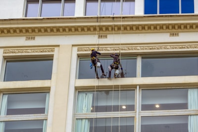 Two workers are suspended on ropes cleaning the windows of a tall building. They are equipped with safety gear, including helmets and harnesses, and are positioned near large glass windows.