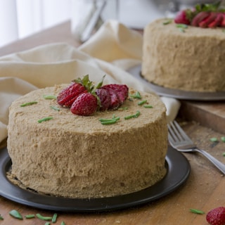 A round cake covered in a crumbly, light brown coating, adorned with fresh strawberries on top. Green sprigs are scattered for garnish. The cake is placed on a dark plate on a wooden surface, accompanied by a fork and a beige cloth in the background.