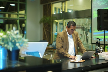 A man in a brown blazer and white shirt is focused on his laptop in a modern, well-lit room. The background features large glass windows and greenery, adding to the contemporary ambiance. On the table, there are a few bottles and a cup on a saucer.