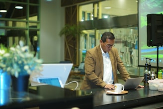 A man in a brown blazer and white shirt is focused on his laptop in a modern, well-lit room. The background features large glass windows and greenery, adding to the contemporary ambiance. On the table, there are a few bottles and a cup on a saucer.