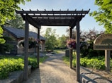A wooden pergola frames a walkway leading to a rustic cabin surrounded by lush greenery. Hanging flower baskets are displayed on the pergola, and a sign reads 'Aurora Community Arboretum'. The scene is bathed in warm sunlight.