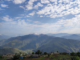 A picturesque view of rolling green hills under a sky dotted with fluffy clouds. Small farmhouses and buildings are nestled at the base of the hills, surrounded by patches of trees and fields. The landscape stretches into the distance with layers of hills creating a sense of depth.