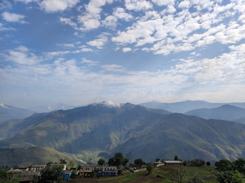 A picturesque view of rolling green hills under a sky dotted with fluffy clouds. Small farmhouses and buildings are nestled at the base of the hills, surrounded by patches of trees and fields. The landscape stretches into the distance with layers of hills creating a sense of depth.