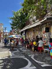 A lively street scene featuring a group of people gathered on a sidewalk. Some individuals are playing drums while others watch or interact around them. The background is adorned with graffiti-covered walls and there is a leafy tree providing some shade. The atmosphere suggests a casual, community-focused event.