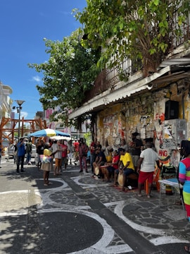 A lively street scene featuring a group of people gathered on a sidewalk. Some individuals are playing drums while others watch or interact around them. The background is adorned with graffiti-covered walls and there is a leafy tree providing some shade. The atmosphere suggests a casual, community-focused event.