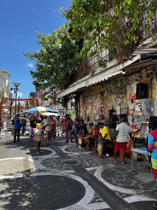 A lively street scene featuring a group of people gathered on a sidewalk. Some individuals are playing drums while others watch or interact around them. The background is adorned with graffiti-covered walls and there is a leafy tree providing some shade. The atmosphere suggests a casual, community-focused event.