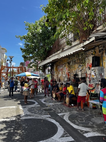 A lively street scene featuring a group of people gathered on a sidewalk. Some individuals are playing drums while others watch or interact around them. The background is adorned with graffiti-covered walls and there is a leafy tree providing some shade. The atmosphere suggests a casual, community-focused event.