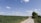 A rural landscape featuring a dirt road leading through green fields under a clear blue sky with scattered clouds. Two large wind turbines are situated in the background, surrounded by open greenery and a solitary tree.