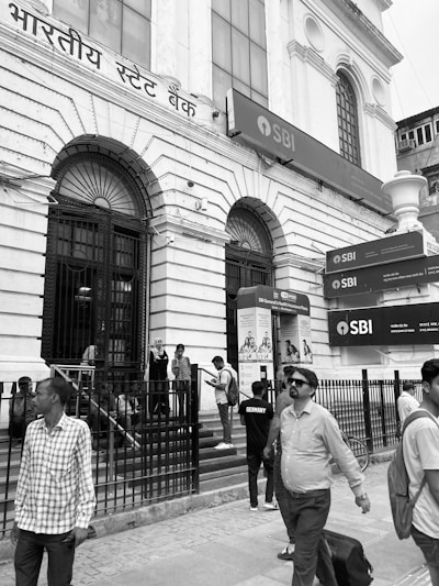 A group of people stands and walks near the entrance of a large bank building with white stone architecture and barred windows. The signage above the entrance is in Hindi, and several SBI logos are visible. The scene captures everyday life outside a busy bank.
