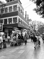 A bustling street scene with people walking past a row of clothing shops. The shops are adorned with hanging garments. A large, intricately designed building is in the background, with architectural features typical of Mughal-style design, including arched windows and ornate decorations. Several tall towers or minarets are visible behind the building. Trees line the street, and a bicycle rickshaw is present.