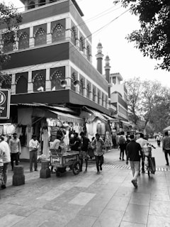 A bustling street scene with people walking past a row of clothing shops. The shops are adorned with hanging garments. A large, intricately designed building is in the background, with architectural features typical of Mughal-style design, including arched windows and ornate decorations. Several tall towers or minarets are visible behind the building. Trees line the street, and a bicycle rickshaw is present.