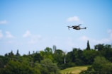 A drone flying in a clear blue sky above a lush, green landscape. Trees and foliage form a backdrop, accentuating the open, airy environment.