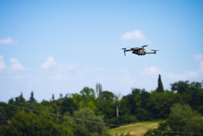 A drone flying in a clear blue sky above a lush, green landscape. Trees and foliage form a backdrop, accentuating the open, airy environment.