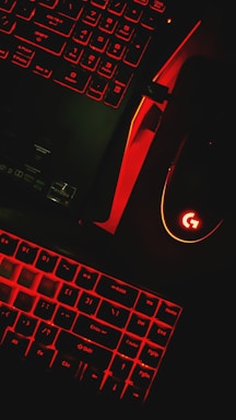 A keyboard and a computer mouse are illuminated by red backlighting. The keys and surrounding areas have a dark background, with the red glow providing stark contrast. The mouse features a prominent glowing logo, and part of a laptop or another keyboard is visible in the upper portion of the image.