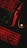 A keyboard and a computer mouse are illuminated by red backlighting. The keys and surrounding areas have a dark background, with the red glow providing stark contrast. The mouse features a prominent glowing logo, and part of a laptop or another keyboard is visible in the upper portion of the image.