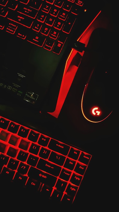 A keyboard and a computer mouse are illuminated by red backlighting. The keys and surrounding areas have a dark background, with the red glow providing stark contrast. The mouse features a prominent glowing logo, and part of a laptop or another keyboard is visible in the upper portion of the image.