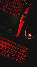 A keyboard and a computer mouse are illuminated by red backlighting. The keys and surrounding areas have a dark background, with the red glow providing stark contrast. The mouse features a prominent glowing logo, and part of a laptop or another keyboard is visible in the upper portion of the image.