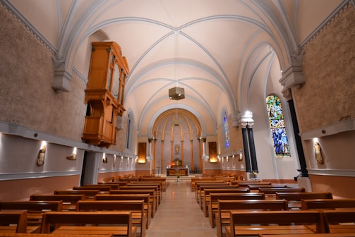 An ornate church interior featuring wooden pews lined in two rows, a high vaulted ceiling with arches, a large pipe organ on the left wall, and a central altar at the far end. Stained glass windows contribute vivid colors to the otherwise warm, subdued lighting. Wall decorations include religious art and icons.