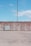 A concrete wall stretches horizontally, with a tall, slim utility pole standing behind it. In front of the wall, there is an electrical box on a slightly raised platform. The scene is set against a clear blue sky with scattered clouds. The ground surface is composed of light-colored paving stones.