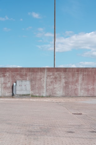 A concrete wall stretches horizontally, with a tall, slim utility pole standing behind it. In front of the wall, there is an electrical box on a slightly raised platform. The scene is set against a clear blue sky with scattered clouds. The ground surface is composed of light-colored paving stones.